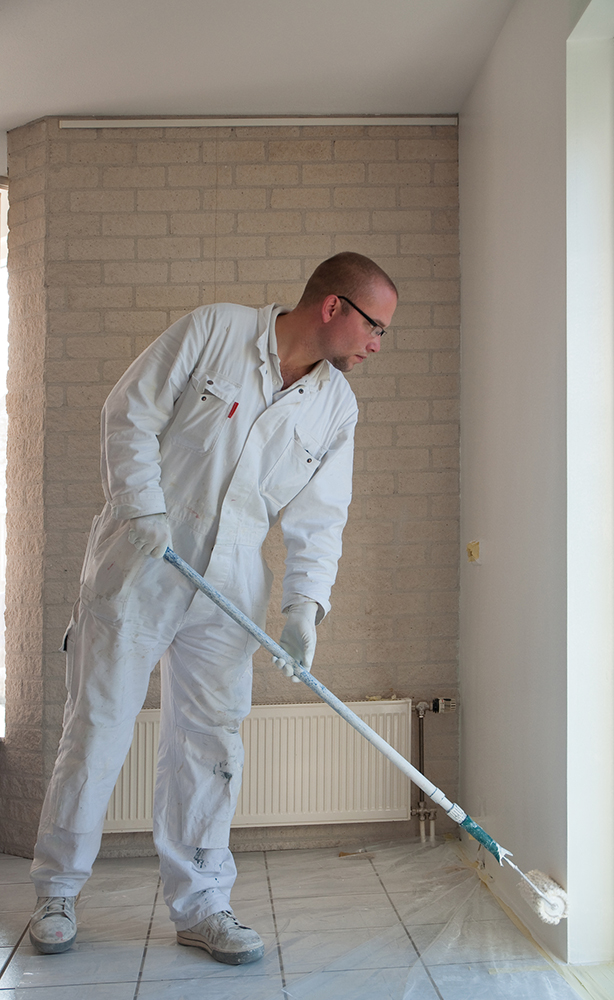 Home decorator painting a wall white with a long roller, standing on plastic to protect the floor tiles